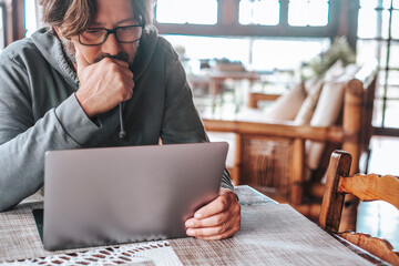 Man working on laptop at home looking thoughtful and focused while using a tablet computer at a rustic wooden table © simona