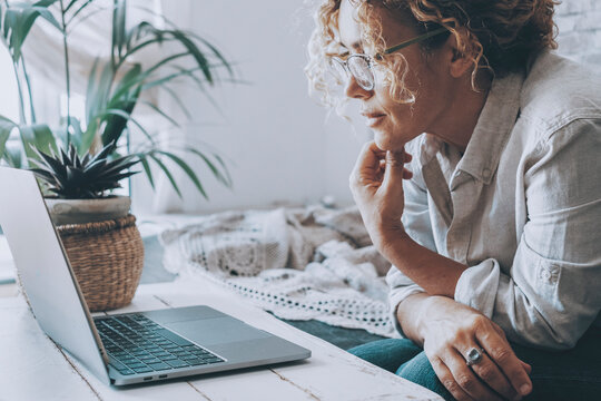 Mature woman wearing glasses working on a laptop at home, leaning on a coffee table beside a potted plant and cozy blanket, focused on remote work and online browsing