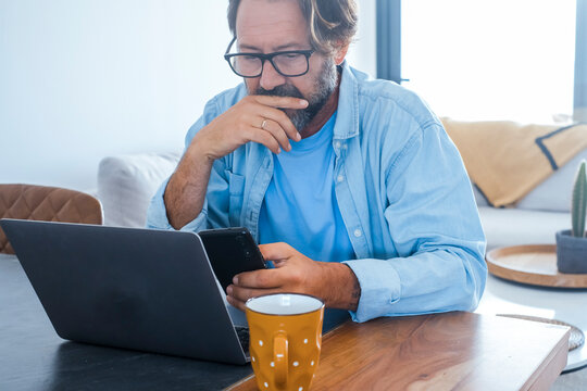 Middle-aged man working from home at a laptop while checking his smartphone, looking thoughtful and focused