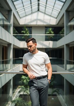 A young man with a beard standing in a multi-story modern office building or shopping mall with a large glass ceiling. He is leaning on a metal railing, wearing a white t-shirt
