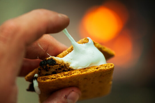 A hand holds a gooey s'mores sandwich of graham crackers, chocolate and melted marshmallows with a campfire in the background