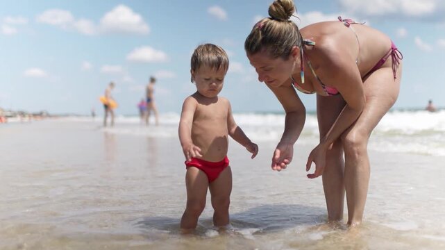 The kid collects shells and pebbles in the sea on a sandy bottom with his mother under the summer sun on vacation