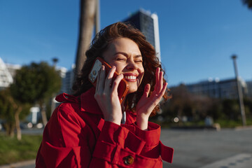 Fototapeta premium Smiling young woman in a red coat talks on the phone in an outdoor urban portrait, a candid phone conversation capturing brunette female confidence in bright afternoon light.