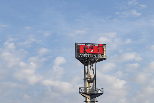 msterdam, Netherlands - 23 May 2026: The iconic red and white RAI Amsterdam logo sign on top of a tall metal advertising tower structure under a bright blue sky with soft white clouds.