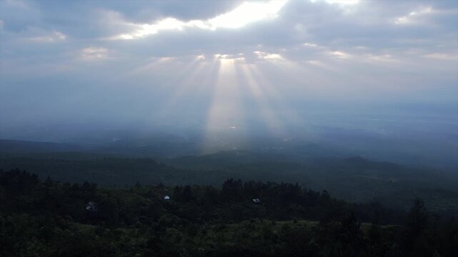 4K aerial view of beautiful dramatic sun rays breaking through clouds over a dark green forest hill.