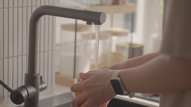 Woman washes orange under running water at kitchen sink. Concept of fruit cleaning and food hygiene