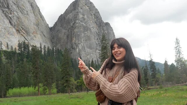 Excited female traveler with a camera smiling and pointing at bridalveil fall, dancing joyfully with el capitan granite monolith and yosemite valley scenery in the background