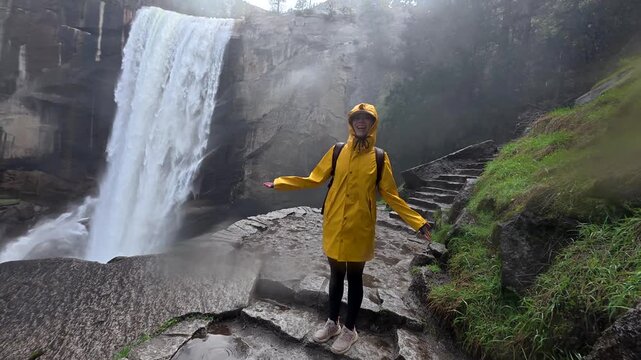Happy young woman wearing a yellow raincoat enjoying the misty spray while hiking on the rocky mist trail next to the powerful vernal fall waterfall in yosemite national park, california