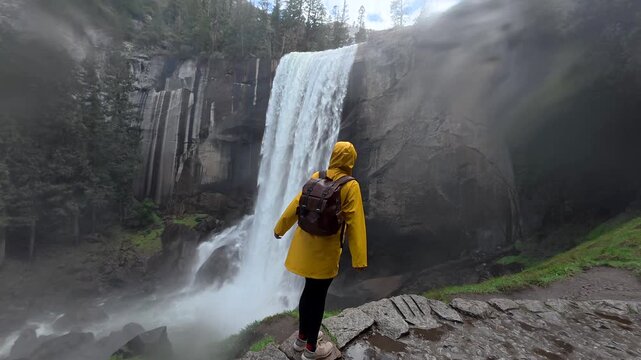Young woman in a yellow raincoat with a backpack stands under the mist of nevada fall in yosemite, getting soaked and soaking in the raw power and scenic mountain view