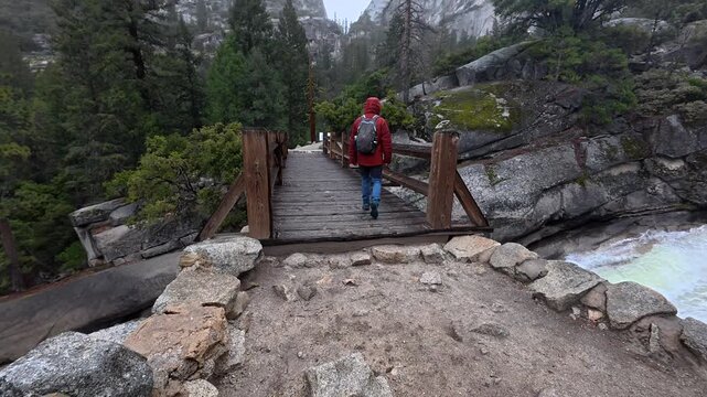 Adventurous man wearing a red rain jacket and carrying a backpack crossing a wooden bridge over a fast flowing river on the famous mist trail in yosemite national park, california