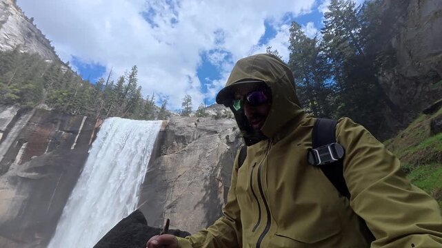Young hiker in a waterproof jacket recording a selfie video while trekking on the wet mist trail, with the powerful vernal fall cascading down granite cliffs in yosemite national park