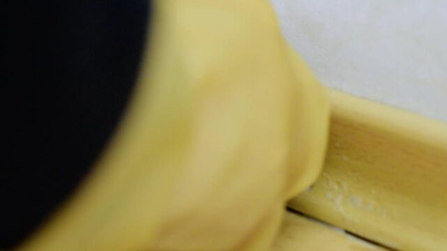 Closeup corner baseboard being scrubbed, yellow glove pressing sponge into joint, removing dirt from floor seam, steady rhythmic motion, focused cleaning task