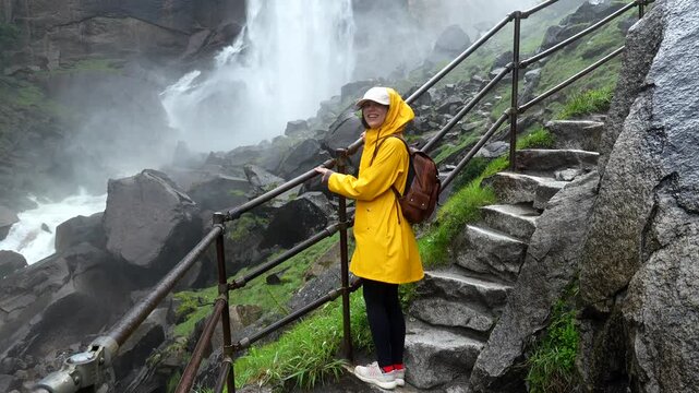 Young smiling woman in a yellow raincoat hiking up the wet stone stairs of the mist trail, enjoying the misty and powerful vernal fall in the majestic yosemite national park, california
