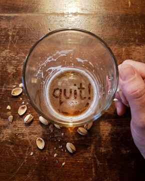 A nearly empty beer glass is held above scattered shells while the thin foam layer and residue at the bottom form the readable word quit!