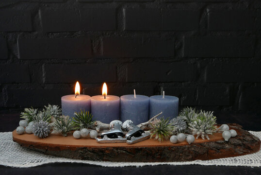 Zweiter Advent: Gesteck mit zwei brennenden blauen Kerze auf einer rustikalen Baumscheibe vor einer schwarzen Backsteinwand mit Textfreiraum.