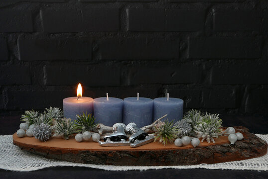  Erster Advent: Gesteck mit einer brennenden blauen Kerze auf einer rustikalen Baumscheibe vor einer schwarzen Backsteinwand mit Textfreiraum.