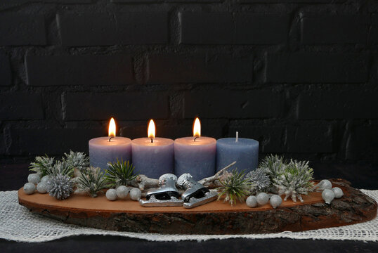 Dritter Advent: Gesteck mit drei brennenden blauen Kerze auf einer rustikalen Baumscheibe vor einer schwarzen Backsteinwand mit Textfreiraum.