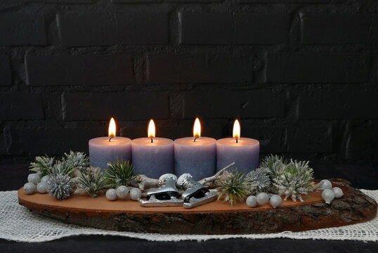 Vierter Advent: Gesteck mit vier brennenden blauen Kerze auf einer rustikalen Baumscheibe vor einer schwarzen Backsteinwand mit Textfreiraum.