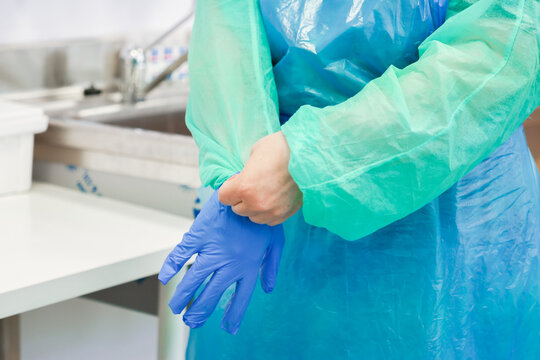 Medical professional wearing a protective blue gown and pulling on latex gloves in a hospital room, establishing clinical safety standards for patient care and infection control