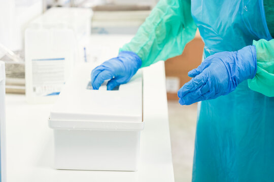 Medical worker in protective apron and blue gloves opens a sterile white plastic container at a clinic table, ensuring hygiene and safety standards in a professional lab environment