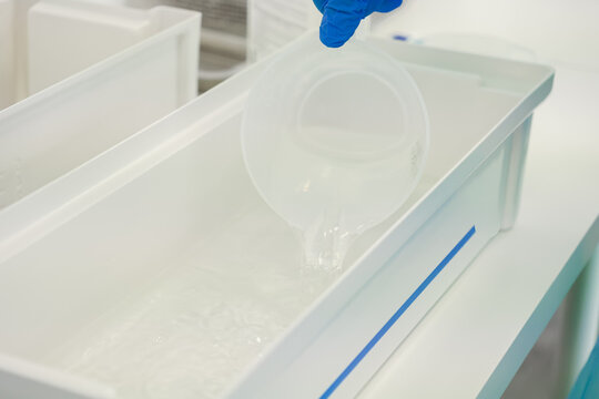 A gloved hand pours clear chemical liquid from a plastic beaker into a white tray in a lab setting, highlighting precise preparation and maintenance of equipment for sterilization