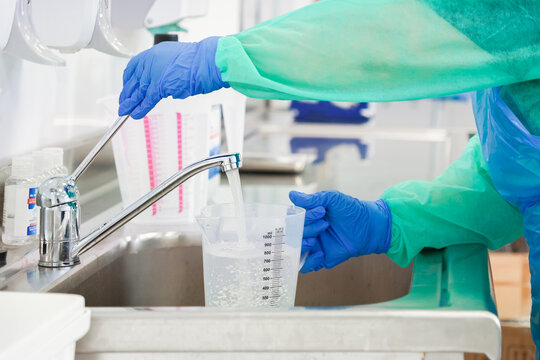 A medical professional in a protective gown and blue gloves fills a plastic graduated cylinder with water at a stainless steel sink for precise scientific laboratory testing