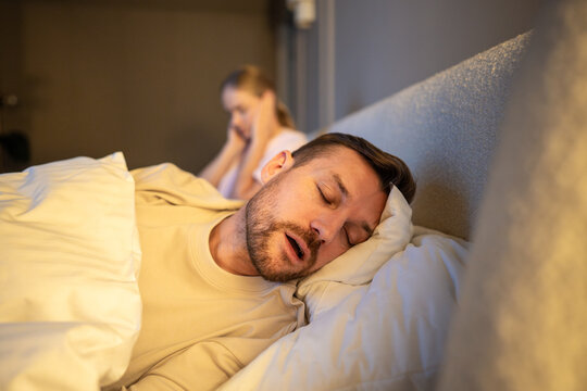 Married couple struggles and relationship stress, tired woman covering ears from noisy snoring sounds of husband sleeping next to her, restless female frustrated by loud disturbing snoring from man
