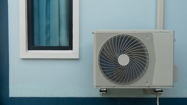 Outdoor air conditioning compressor unit mounted on the exterior wall of a light blue building. A glass window pane is visible next to the ventilation equipment.