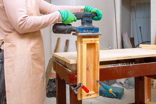 Woman carpenter using an orbital sander to finish a wooden piece on a workbench. Professional woodworker sanding lumber in a workshop. Home improvement and renovation carpentering project