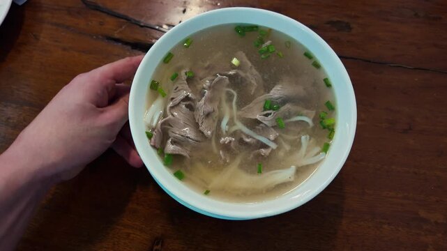 Server's hands placing a steaming bowl of traditional vietnamese beef noodle soup, pho bo, onto a dark wooden table in an authentic asian restaurant for a savory and hearty dining experience