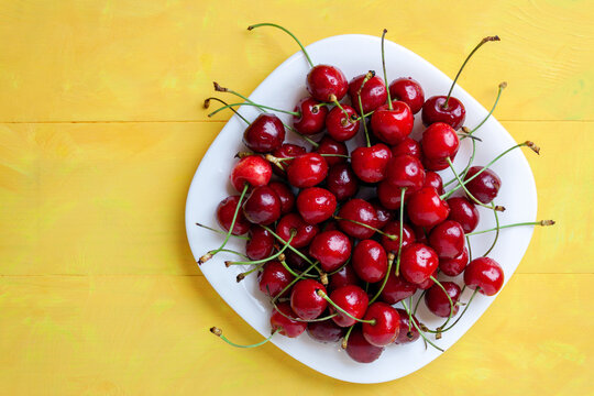Fresh red cherries with green stems served on a white ceramic plate over a yellow wooden table. sweet summer berry,