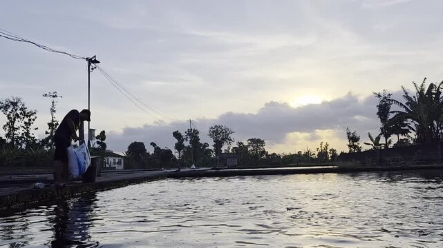 Wide shot of an Asian male farmer silhouette feeding fish in a large pond during sunset or sunrise. Aquaculture, traditional fish farming, and rural agriculture lifestyle concept.
