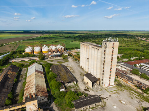 Aerial view of a tall concrete grain elevator and industrial warehouses with rusty roofs surrounded by green fields in Balchik, Dobrich Province, Bulgaria.