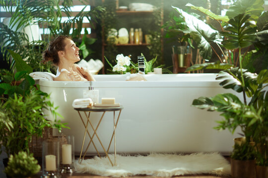 A smiling woman with a bun enjoys a relaxing bath in a white freestanding tub, enveloped by lush green plants and natural light, creating a serene and luxurious home spa retreat.