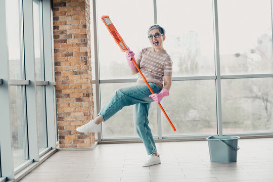 Joyful woman dancing while mopping near large windows wearing pink gloves and casual jeans during a bright home cleaning session