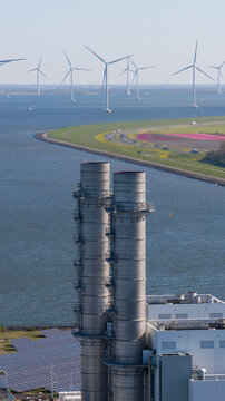Aerial view of the Maxima power plant with tall industrial chimneys, solar panels, and offshore wind turbines near colorful tulip fields in Lelystad, Flevoland, Netherlands.