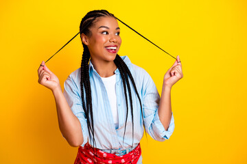 Young woman with braids pulls hair ties against bright yellow background smiling for fashion...