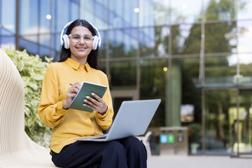 Young female student smiling on a campus bench, wearing headphones, typing on a laptop and jotting...