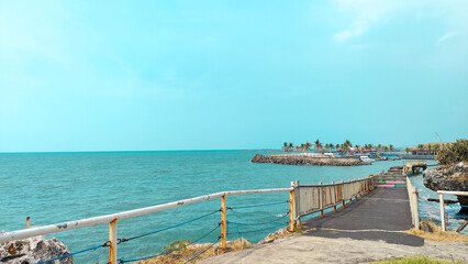 Fototapeta premium A rustic coastal walkway with white railings leading toward a tropical island with palm trees, surrounded by calm turquoise ocean waters under a clear sky in a peaceful summer setting