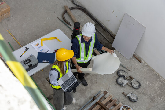 Professional engineer and worker stand in construction site holding blueprint wearing hardhat inside an uncompleted building displaying focused teamwork on development project
