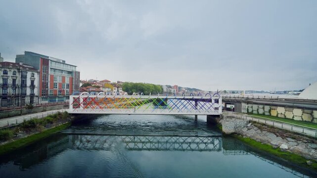 Modern white pedestrian bridge decorated with a vibrant rainbow art installation, spanning a tranquil river in an urban setting with its beautiful reflection appearing on the water surface