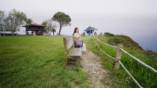 Woman sitting on a wooden bench in a green meadow taking a selfie with her smartphone. Scenic coastal location with a traditional horreo and a small chapel in the background on a cloudy day