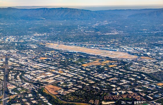 Aerial panorama of San Jose Mineta International Airport in California, showing its runways surrounded by a grid of industrial buildings, residential areas, and the Santa Clara Valley foothills