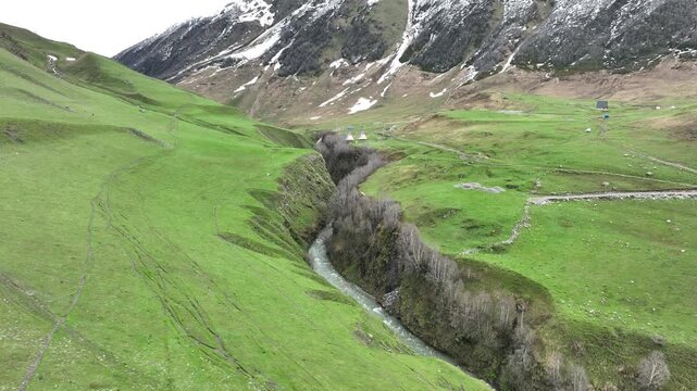High-angle aerial view capturing a clean, rushing mountain creek flowing from a melting snow patch down a vibrant green alpine valley with rocky terrain and small wildflowers in Ushguli, Georgia
