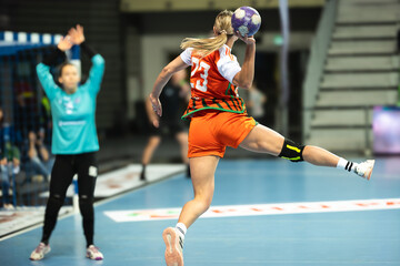 Naklejka premium A female handball player in an orange jersey jumps to throw the ball towards the goal defended by a goalkeeper in blue.