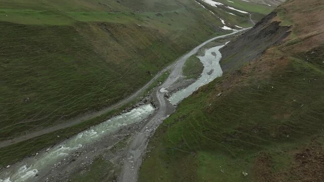 High-angle aerial view capturing a clean, rushing mountain creek flowing from a melting snow patch down a vibrant green alpine valley with rocky terrain and small wildflowers in Ushguli, Georgia