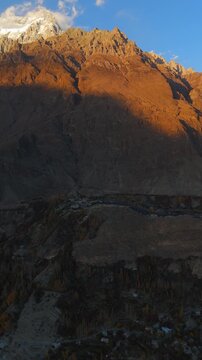 Aerial view of the Hunza valley with golden hour light hitting the rocky mountain peaks and snow-capped summits in Hunza Nagar, Gilgit Baltistan, Pakistan.