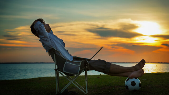 Relaxed man sitting on camping chair with feet on soccer ball near lake working with laptop during sunset for digital nomad and remote work concept