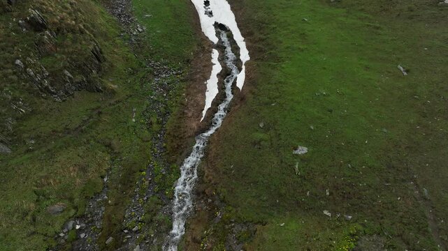 High-angle aerial view capturing a clean, rushing mountain creek flowing from a melting snow patch down a vibrant green alpine valley with rocky terrain and small wildflowers in Ushguli, Georgia