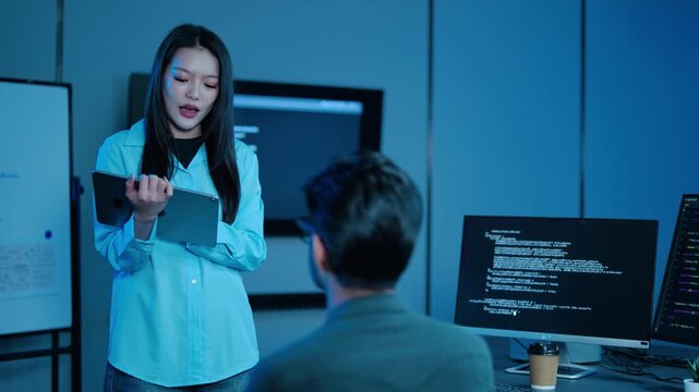 Asian female IT project manager discussing software code with male programmer at night in dark blue modern tech office. Startup team working late on computer programming, analyzing data together.